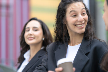 Obraz premium A diverse team of three Hispanic businesswomen in dark suits, laughing with genuine warmth, listen as a teammate explains her thinking with one raising her cup to drink.