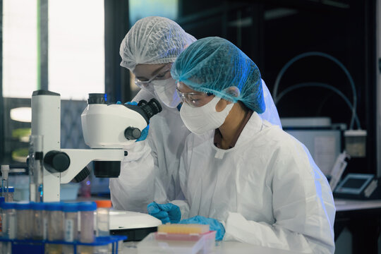 Two Scientists in Protective Gear Examining Sample Under Stereo Microscope in Modern Laboratory