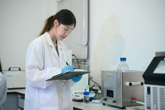 Young Asian Female Scientist Writing Notes on Clipboard in Modern Laboratory with Ultrasonic Cleaner Equipment