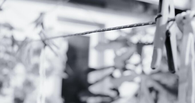 Black and white photo of three clothespins hanging on a wire clothesline outdoors. Soft blurred background with garden plants and a fence. Vintage film grain style. Everyday rustic scene.