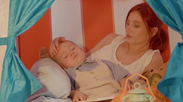 A mother lovingly caresses her son in a colorful tent as she tells him delightful stories. The sweet interaction helps the little boy drift off to sleep peacefully.