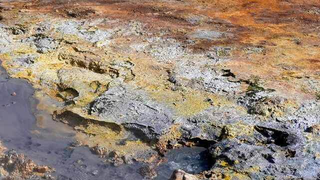 Close-up of colorful geothermal ground at Kerlingarfjoll in Iceland, with sulfur-yellow and rust-orange mineral crusts surrounding a gray mud pool.