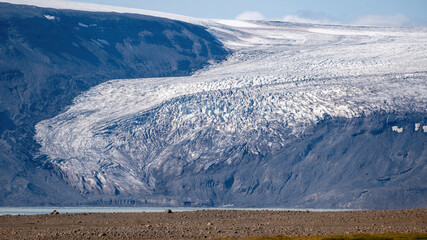 Massive glacier tongue flows down dark volcanic slopes toward a barren gravel plain in the Icelandic Highlands, showing visible effects of climate change. © Asanee