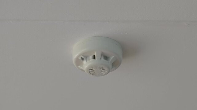 Smoke detector mounted on a white ceiling with a red led light blinking, signaling that the fire alarm system is active and working properly