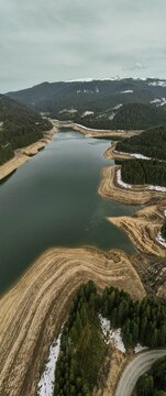 Wide view of a winding river surrounded by forested mountains with snow patches and dry land creating natural contours at a reservoir in Romania during cool weather