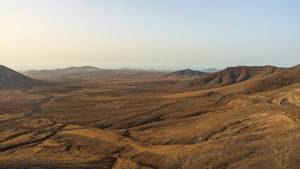 Fototapeta premium A sweeping view from Mirador de la Muda overlooks a vast arid valley and winding roads leading toward distant volcanic hills, Fuerteventura, Canary Islands, Spain.