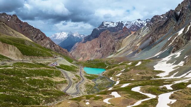 Winding highway curves through rugged alpine mountains, beside a turquoise lake with snow patches under bright daytime sunlight.