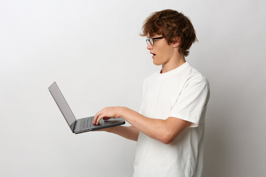 Young man in glasses and white t-shirt holds a laptop and reacts with surprise, mouth open. Side view on a gray background. Concept for news, technology or online sales.