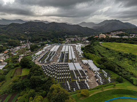 Aerial view of a large truck storage park filled with rows of white trucks, surrounded by lush green hills and trees under a cloudy sky in Atsugi, Kanagawa, Japan.