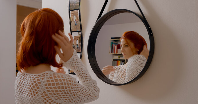Young redhead carefully adjusting her hair while checking her reflection, standing in bright, minimalist home interior during morning routine