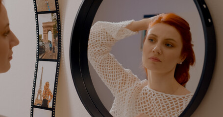 Young redhead woman stands in front of a mirror, visibly displeased as she tries to fix her hairstyle. She expresses frustration with her appearance in her bedroom. © Binzari