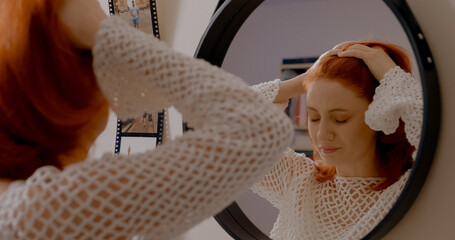 A redhead woman stands in front of a mirror, massaging her temples as she battles a headache. She prepares to leave her home while feeling the effects of migraines. © Binzari