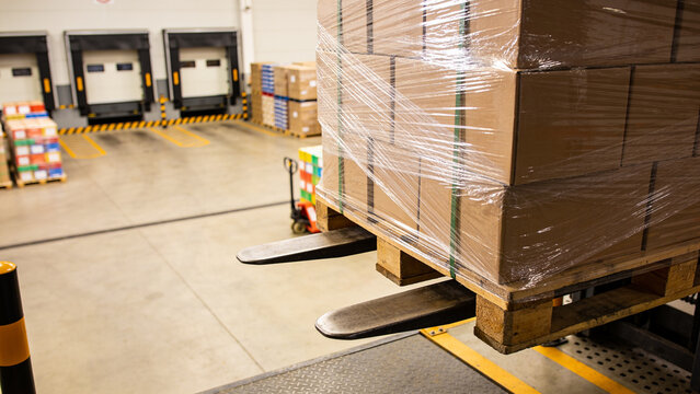 Stacked cardboard pallet on forklift forks in a warehouse loading dock, shrink wrapped and ready for loading or transport in an industrial logistics setting.