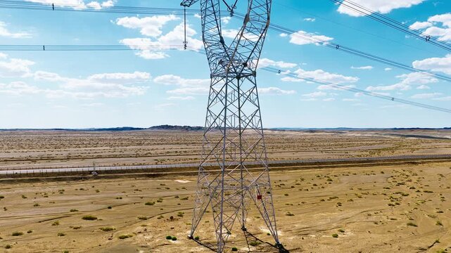 High-voltage electricity pylon stands on dry desert terrain beside an asphalt road under bright sunny skies with scattered cumulus clouds.