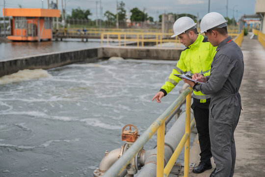 Engineer and technician inspect a wastewater treatment basin and record operational data beside industrial pipelines. Concept of water quality, process control, utilities, and engineering.