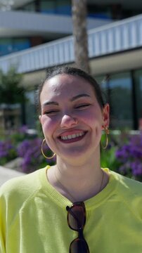 Vertical close portrait of a cheerful young White woman walking toward the camera and laughing in bright outdoor sunlight.