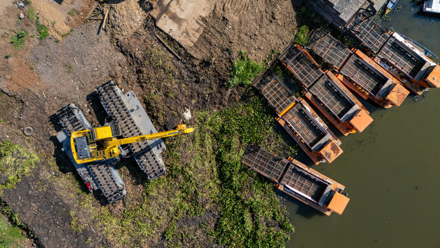 Aerial view of an excavator mounted on a floating platform working near a riverbank with several moored barges in Salatiga, Central Java, Indonesia.