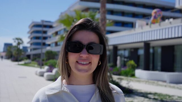 A happy young White woman wearing sunglasses walks toward the camera and smiles on a sunny modern city promenade.