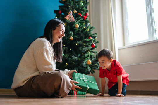 Mother and son open Christmas gift beside tree at home
