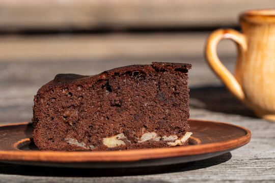 Chocolate cake slice on plate, closeup. Slice of homemade chocolate brownie cake with apples on clay plate, with ceramic cup blurred in the background on wooden table
