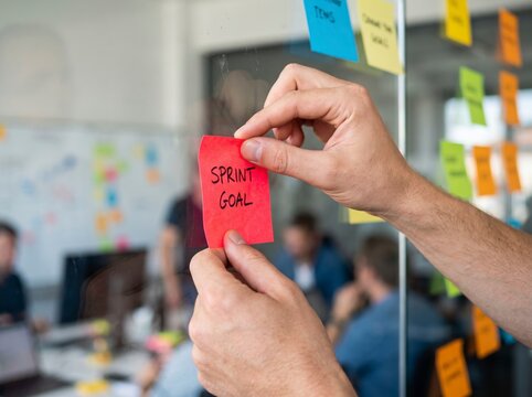 Close-up of hands attaching a red post-it note labeled Sprint Goal onto a glass board during an agile scrum team planning session in a modern tech office.