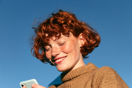 Cheerful Gen Z woman with smartphone under blue sky