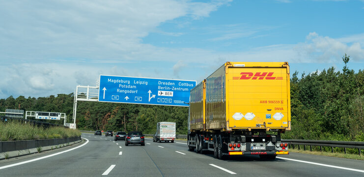 Germany - Aug 22, 2026: Wide view of a yellow DHL road train approaching a blue sign gantry on a motorway with dark cars in the middle lane and pine woodland along the far verge