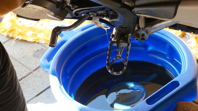 Used oil draining from motorcycle engine into blue plastic basin. Motorbike maintenance and oil change process. DIY vehicle repair and regular service concept.
