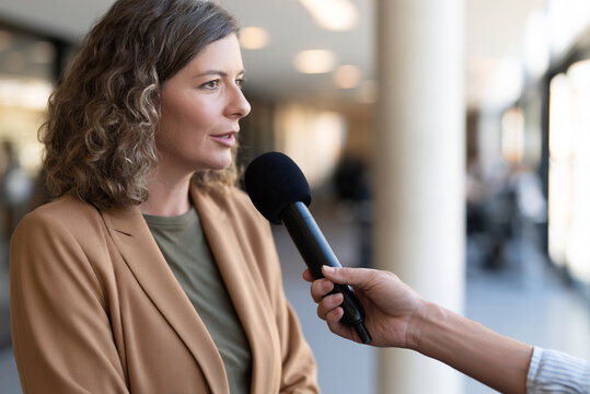 Female journalist holding microphone while interviewing business professional, focus on interviewee, bright contemporary office, news media concept.