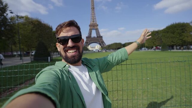 Cheerful caucasian man making a selfie video in Paris with the Eiffel Tower in the background, enjoying a sunny sightseeing trip, looking at camera and waving smiling.