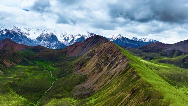 Green mountain ridges and deep valleys extend toward snow-capped alpine peaks under a dynamic cloudy sky illuminated by soft daylight.