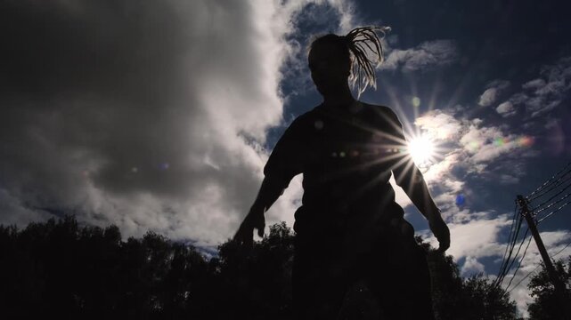 A silhouette of a man with long dreadlocks playing basketball, bouncing the ball on the floor against the backdrop of the sky and bright sun