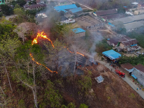 Aerial view of a forest fire spreading through dry vegetation near a residential area with a fire truck and people on the road in Indonesia.