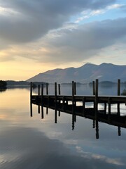 Fototapeta premium Wooden pier extending into a calm mountain lake at sunset, with dramatic clouds and beautiful reflections on the peaceful water.