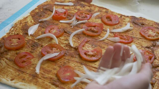 Tomato paste covers rolled dough beside chopped tomato pieces while hand adds fresh onion. Simple vegetable combination creates balanced flavor, natural color and appetizing pizza preparation
