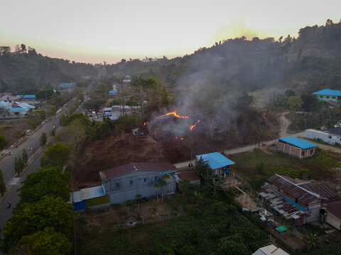 Aerial view of a brush fire burning on a hillside near Waisai town, with smoke rising above residential houses, roads, and lush vegetation in Raja Ampat, Southwest Papua, Indonesia.