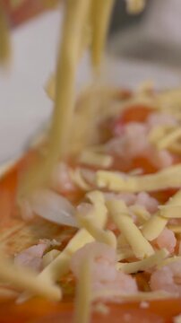 Hand scatters hard grated cheese pieces across pizza dough with cooked shrimp, sweet pepper and tomato. Colorful ingredients create rustic visual contrast and suggest homemade culinary preparation