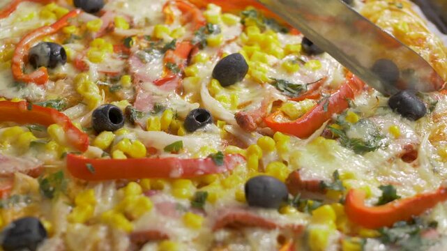 Clumsy cutting pizza into slices on large tray by knife, perfect for groups. Party gatherings, celebrations, and casual events. Corn, red pepper, ham, olive across dough beneath smooth melted cheese