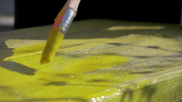Cottage decor receives attention. Yellow coat goes on wooden board with help of brush. Female hand defines edges. Quiet space supports mindful task. Wooden texture shows under paint
