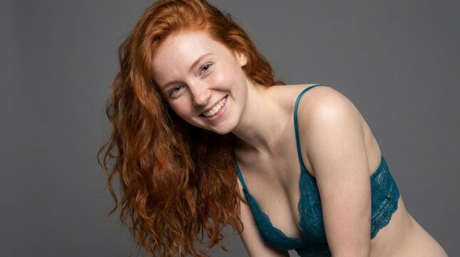 Smiling young woman with long red hair and freckles wearing a teal lace bralette against a gray background, looking at the camera, concept of natural beauty and confidence
