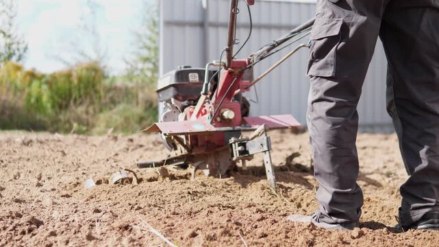 Low-angle rural farming scene shows worker in dark workwear guiding compact tilling machine through dry soil, dirt scattering in foreground near blurred shed and surrounding vegetation under bright