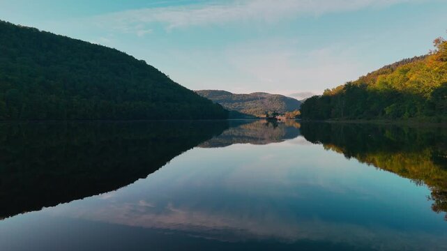Calm water slow dolly drone view at Sherman Reservoir Boat Launch, Vermont, USA, with forested mountains, tree reflections and clear sky. Sunny weather. Peaceful nature and boating recreation. 4K.