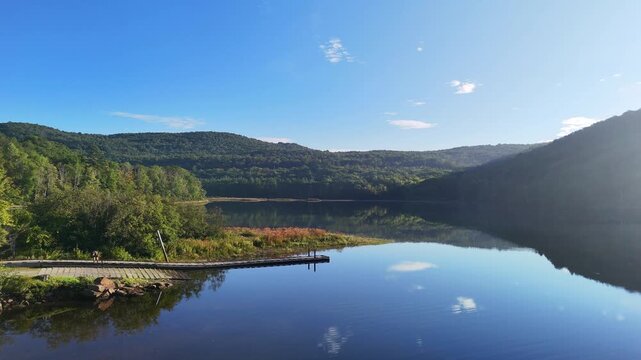 Peaceful nature and boating recreation. Scenic water left to right drone view at Sherman Reservoir Boat Launch, Vermont, USA, with forested mountains, tree reflections and clear sky. 4K.
