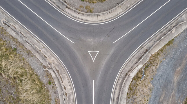 An overhead drone shot captures a grey asphalt Y-junction road with clear white lane markings and dry grass and gravel surroundings.