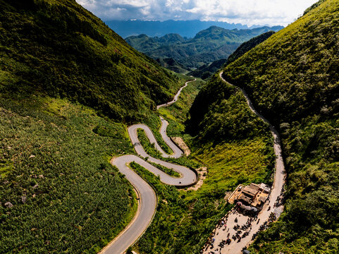 Aerial view of the winding Tham Ma Pass road on the Ha Giang Loop, surrounded by lush green mountains and steep valleys, with a small roadside stop in Dong Van, Tuyen Quang, Vietnam.