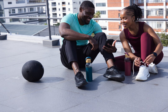 African friends wearing workout clothes tying laces on rooftop and checking phone near med ball