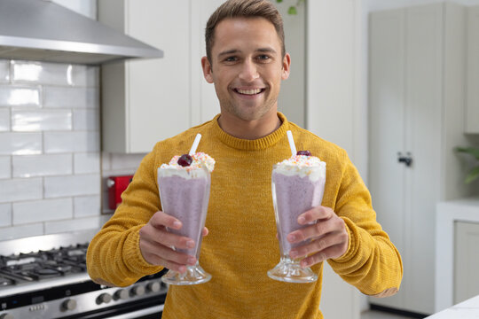 Man in late twenties wearing mustard sweater standing in kitchen holding two purple milkshakes