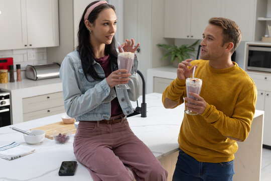Couple sharing milkshakes while woman sitting on marble island and man standing by sink