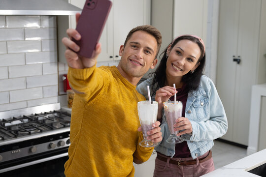 Couple taking selfie in kitchen wearing mustard sweater denim jacket holding phone and milkshakes