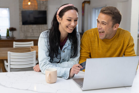 Couple leaning on white marble countertop, looking at laptop, holding credit card, mug nearby